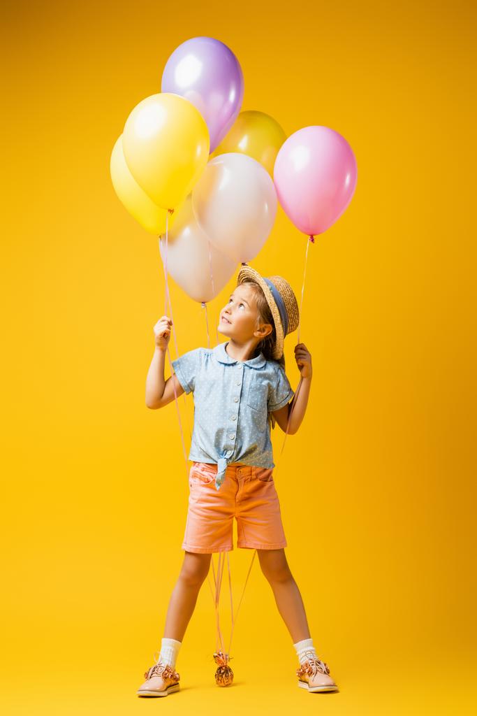 полная длина счастливого ребенка в соломенной шляпе глядя на воздушные шары на желтый - Фото, изображение
