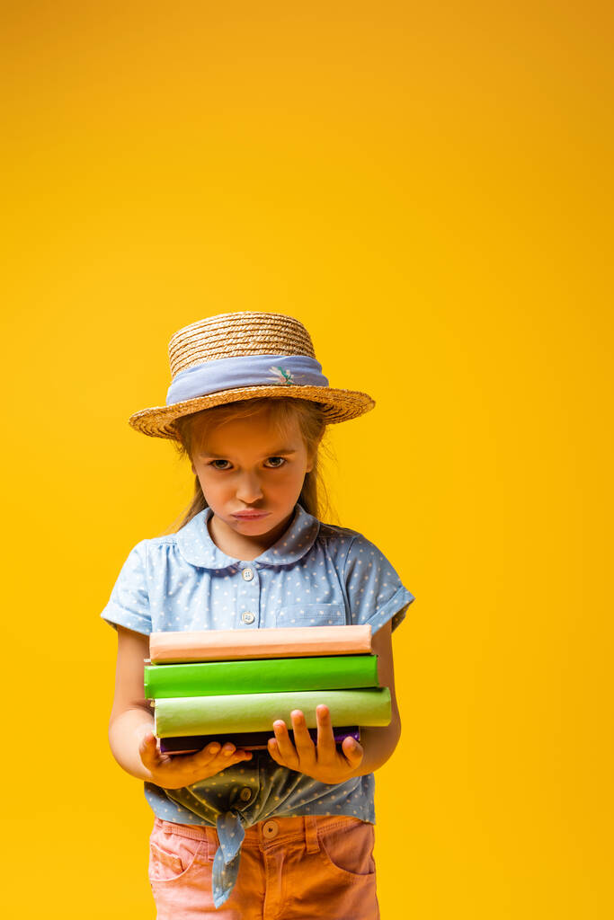 λυπημένο παιδί με ψάθινο καπέλο που κρατάει βιβλία απομονωμένα στο κίτρινο - Φωτογραφία, εικόνα