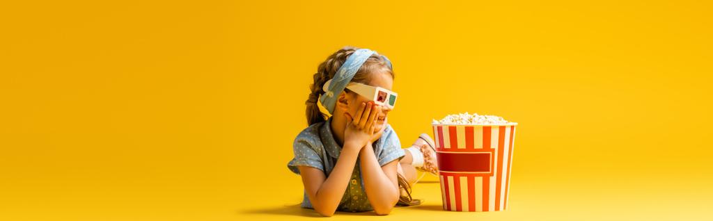 happy kid in 3d glasses lying near popcorn bucket on yellow, banner - Photo, Image