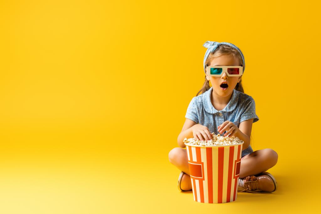 yllättynyt lapsi 3D-lasit istuu ristissä jalat lähellä popcorn ämpäri ja katsella elokuvaa keltainen  - Valokuva, kuva