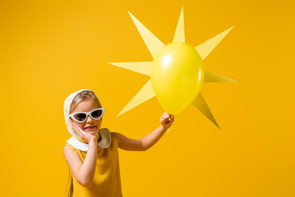 boldog lány fejkendőben és napszemüvegben közel dekoratív nap léggömbbel elszigetelt sárga - Fotó, kép