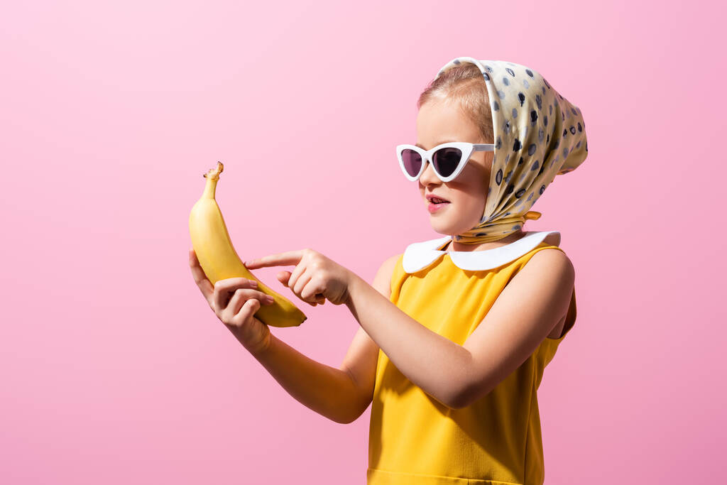 счастливая девушка в платке и солнцезащитных очках указывая пальцем на банан, изолированный на розовый - Фото, изображение