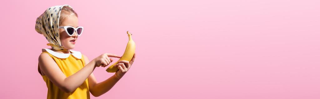 menina feliz em lenço de cabeça e óculos de sol apontando com o dedo para banana isolada em rosa, banner - Foto, Imagem