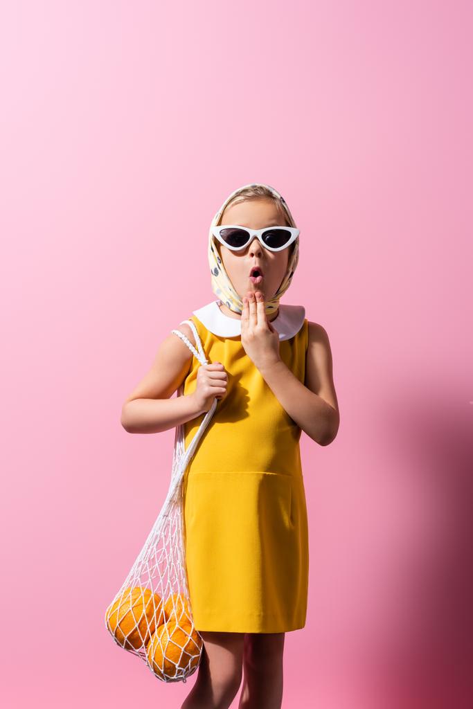 потрясенная девушка в платке и солнцезащитных очках держит многоразовый мешок с апельсинами на розовом - Фото, изображение