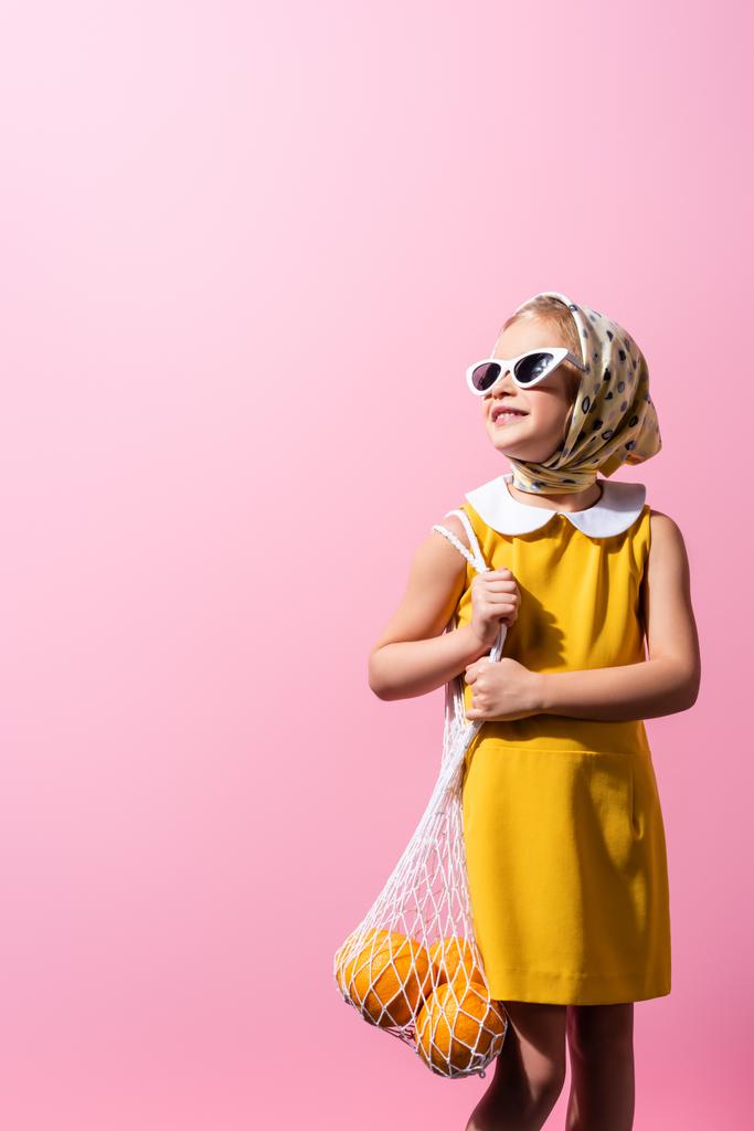 iloinen tyttö huivi ja aurinkolasit tilalla uudelleenkäytettäviä merkkijono laukku appelsiinit eristetty vaaleanpunainen - Valokuva, kuva