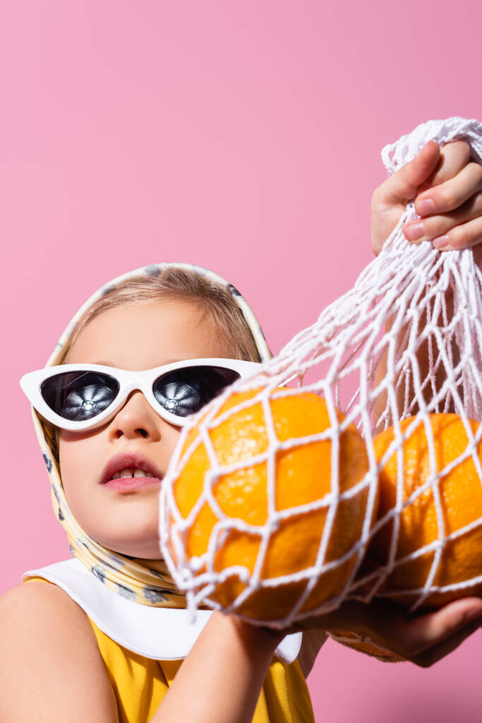 девушка в платке и солнцезащитных очках держа многоразовый мешок с апельсинами на размытом переднем плане изолированы на розовый - Фото, изображение