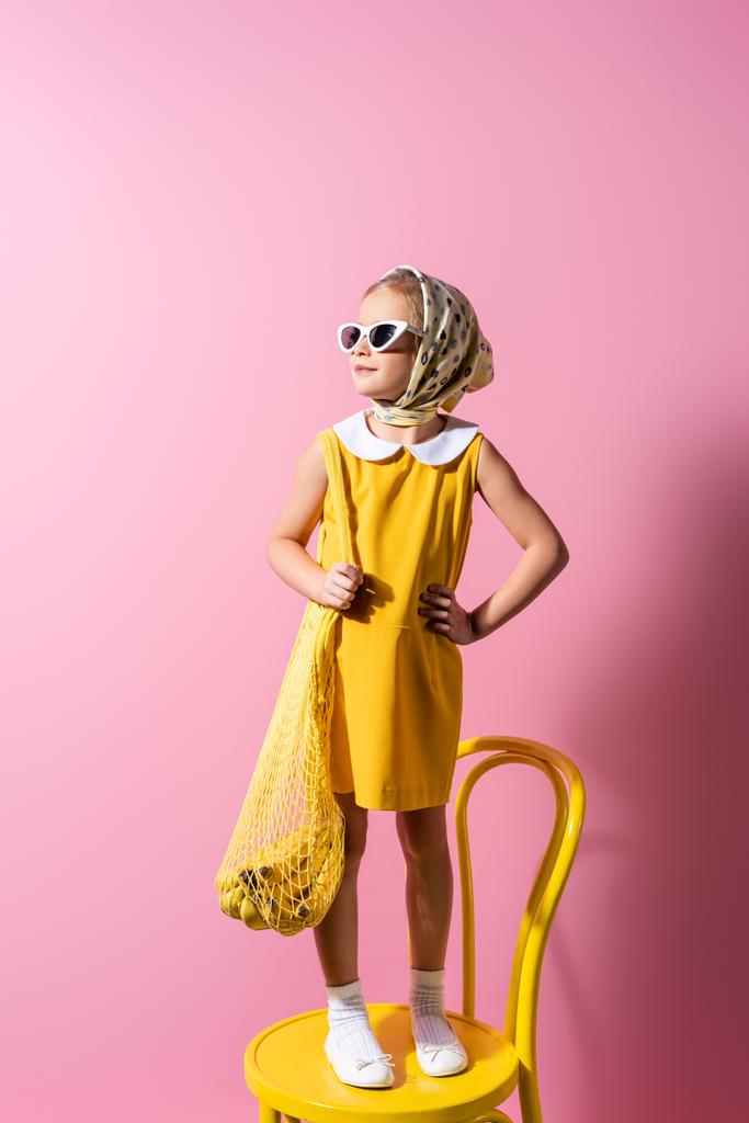 cheerful girl in headscarf and sunglasses holding reusable string bag with bananas while standing on yellow chair on pink - Photo, Image