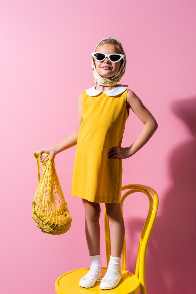 szczęśliwa dziewczyna w chustce i okularach przeciwsłonecznych gospodarstwa wielokrotnego użytku stringi torba z bananami stojąc z ręką na biodrze na różowym - Zdjęcie, obraz