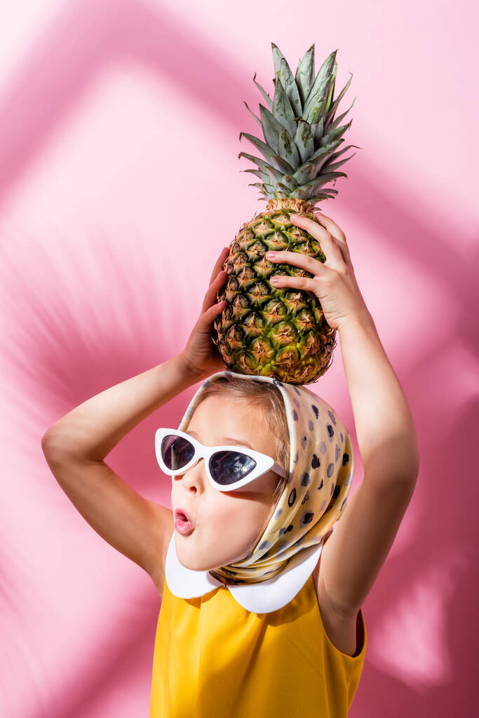 Schockiertes Mädchen mit Kopftuch und Sonnenbrille hält Ananas über dem Kopf auf rosa  - Foto, Bild