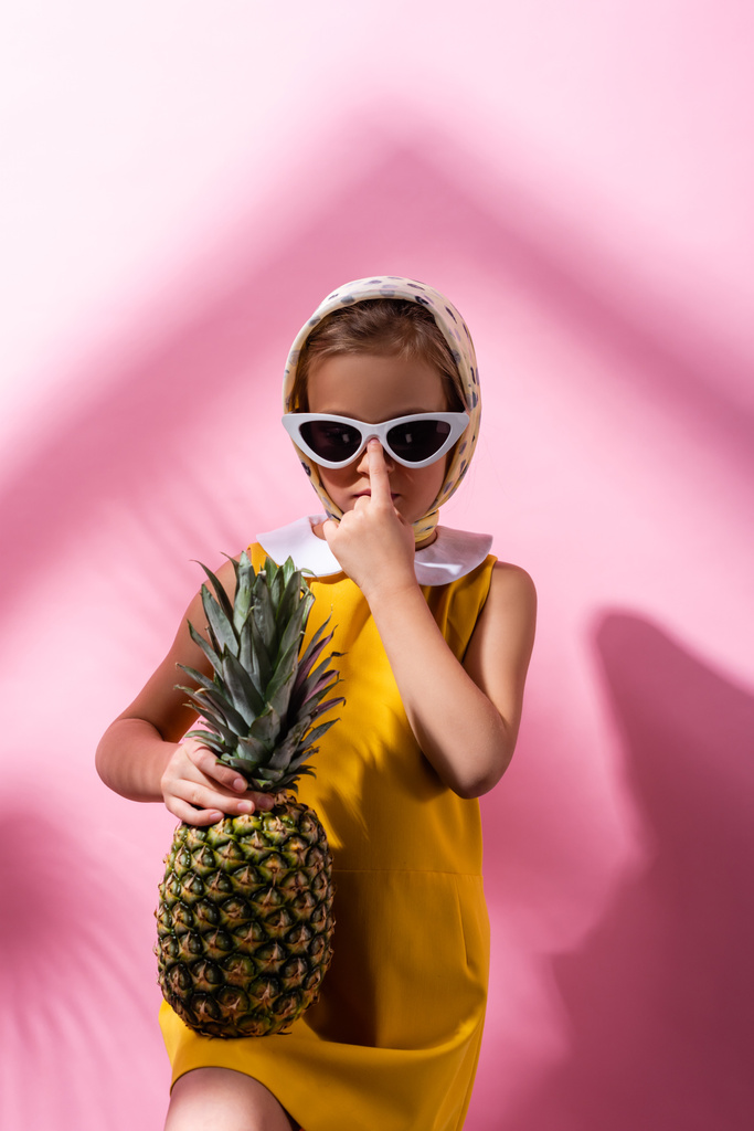 menina em lenço de cabeça ajustando óculos de sol enquanto segurando abacaxi em rosa  - Foto, Imagem