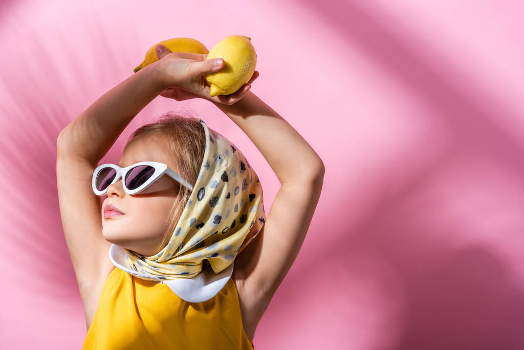 stylisches Mädchen mit Kopftuch und Sonnenbrille, Zitronen über dem Kopf auf rosa  - Foto, Bild