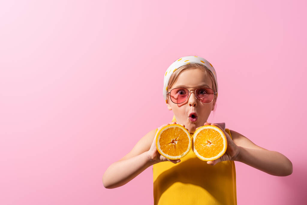 fille choquée en foulard et lunettes de soleil tenant des moitiés orange sur rose  - Photo, image