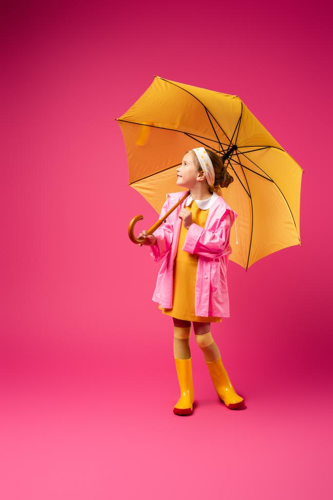 volledige lengte van positief meisje in regenjas staande onder gele paraplu op karmozijn - Foto, afbeelding