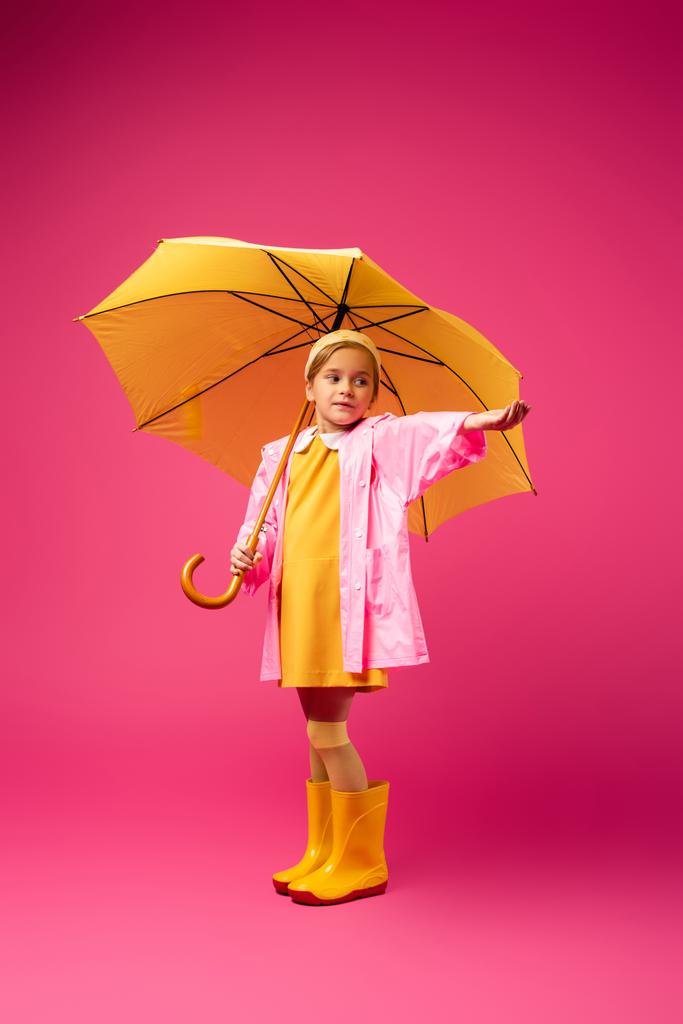 Mädchen in Regenmantel und Gummistiefeln stehen unter gelbem Regenschirm auf purpurrotem Grund - Foto, Bild
