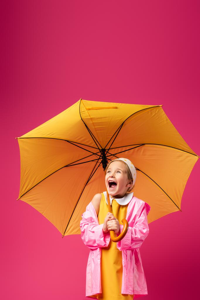 verbaasd meisje in regenjas staan onder paraplu op karmozijn - Foto, afbeelding