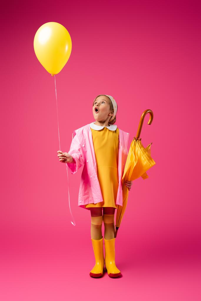πλήρες μήκος του έκπληκτος κορίτσι με αδιάβροχο και μπότες από καουτσούκ κρατώντας μπαλόνι και κίτρινη ομπρέλα σε πορφυρό - Φωτογραφία, εικόνα