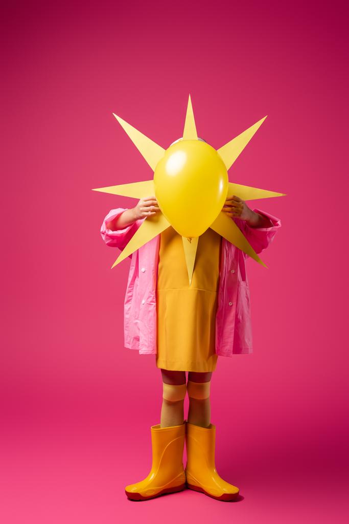 longitud completa de la muchacha en impermeable y botas de goma cubriendo la cara mientras sostiene el sol decorativo con globo en carmesí - Foto, Imagen