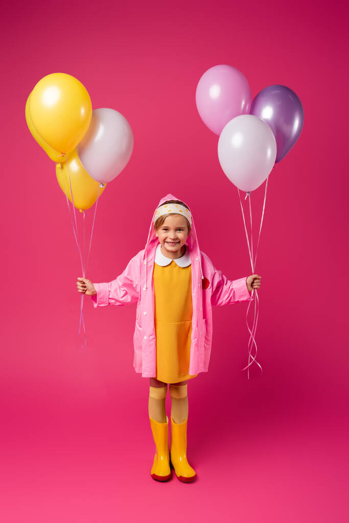 full length of happy girl in raincoat and rubber boots holding balloons on crimson - Photo, Image