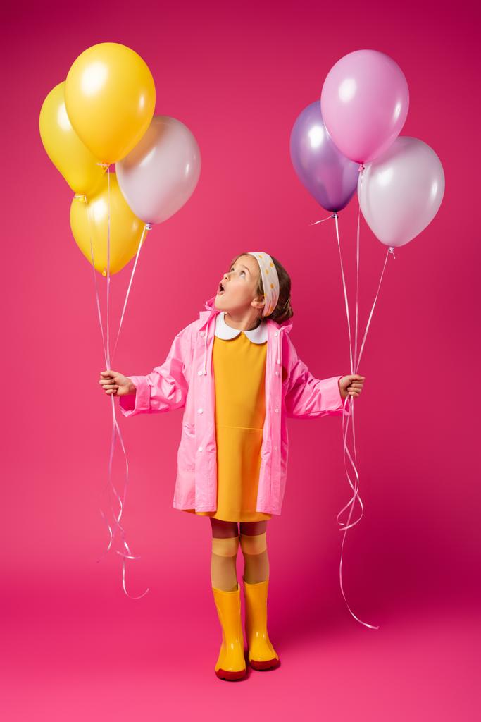 in voller Länge staunende Mädchen in Regenmantel und Gummistiefeln, die Luftballons auf purpurrotem Grund halten - Foto, Bild