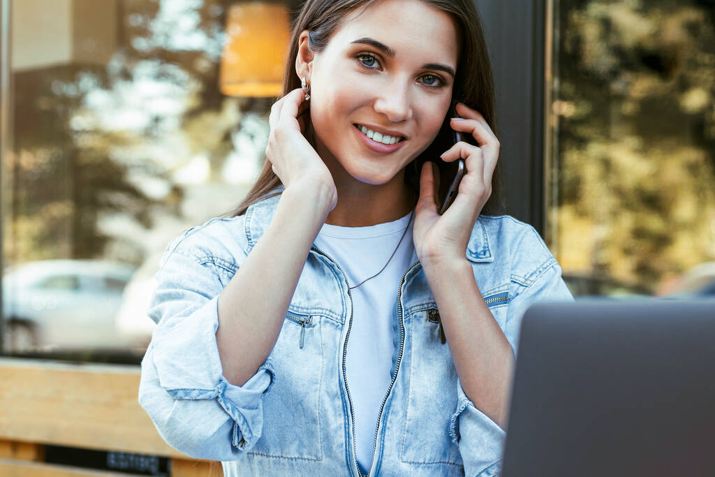 Jonge zakenvrouw werkt op een open terras thuis, zitten in de voorkant van de laptop, met de smartphone in haar hand.  - Foto, afbeelding