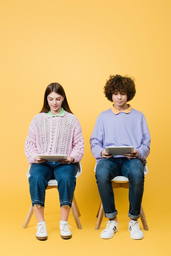 Nastolatki korzystające z tabletów cyfrowych na krzesłach na żółtym tle - Zdjęcie, obraz