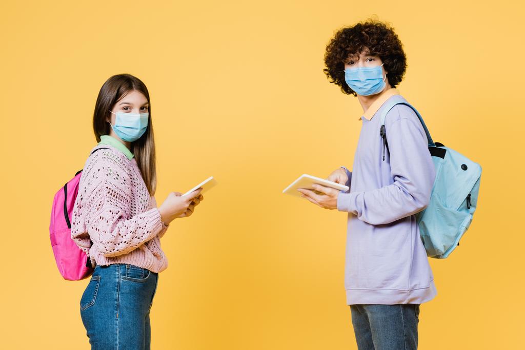Teini-ikäiset, joilla on reput ja lääketieteelliset naamarit käyttäen keltaisella eristettyjä digitaalisia tabletteja - Valokuva, kuva