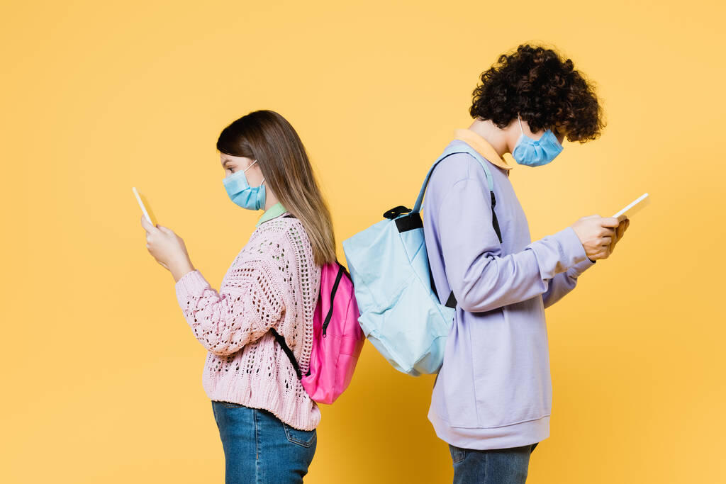 Side view of teen friends with backpacks and medical masks using digital tablets on yellow background - Photo, Image