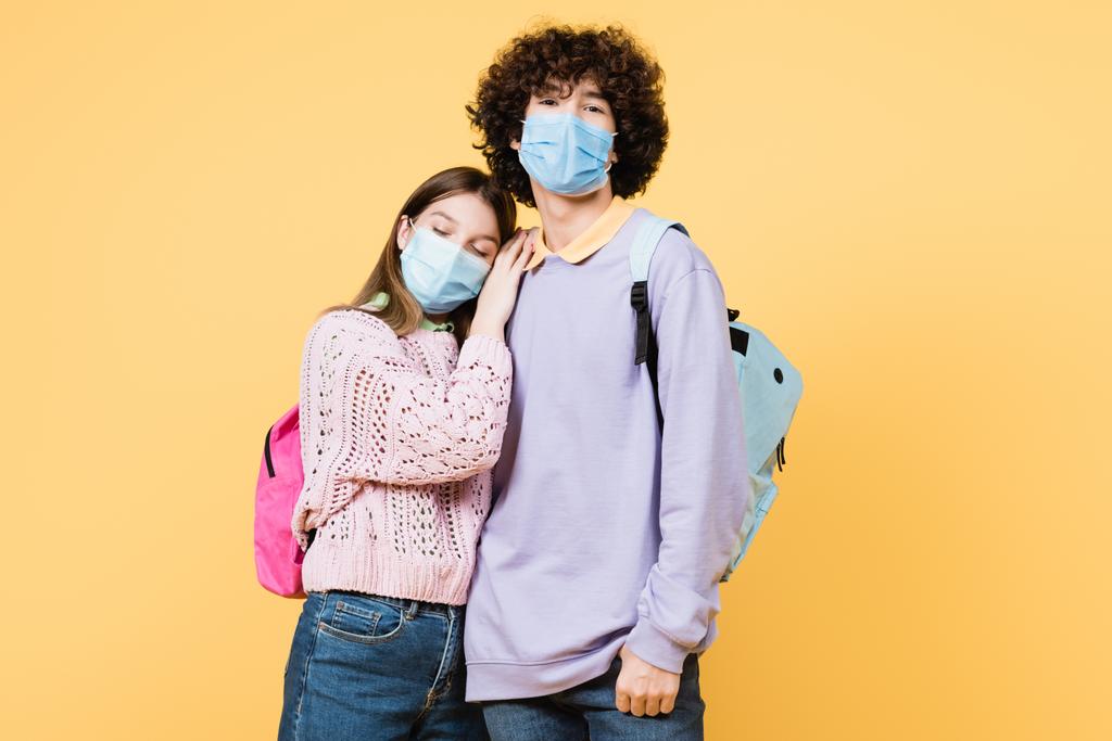 tiener meisje in medisch masker omarmen vriend met rugzak geïsoleerd op geel - Foto, afbeelding