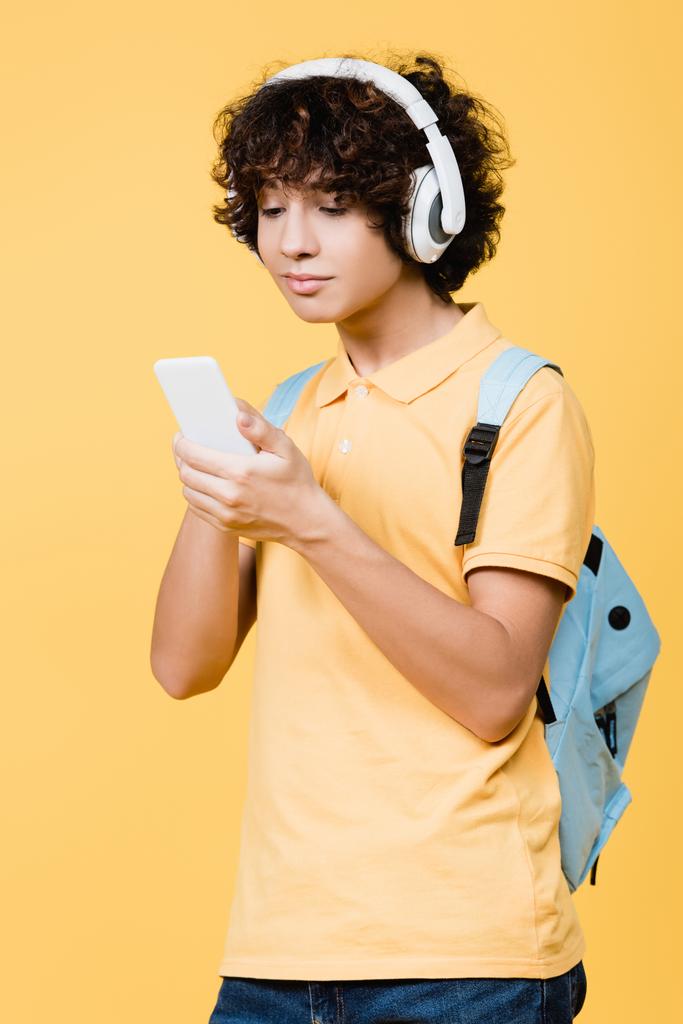 adolescent garçon écoute de la musique dans casque et en utilisant téléphone cellulaire isolé sur jaune - Photo, image