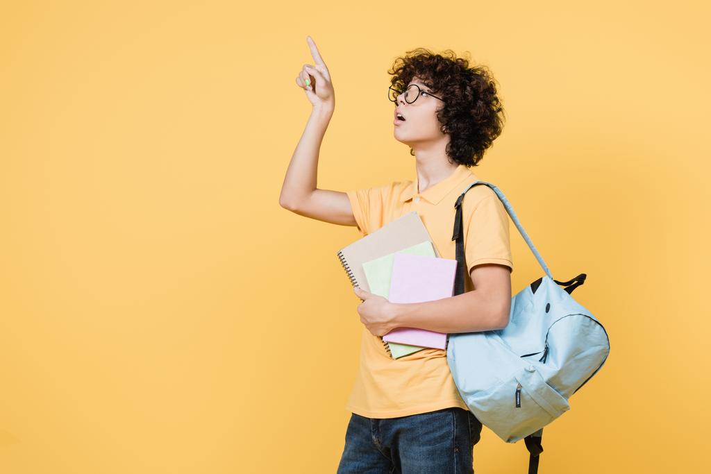Lockiger Schuljunge mit Rucksack und Notizbüchern, die mit dem Finger auf gelb zeigen - Foto, Bild