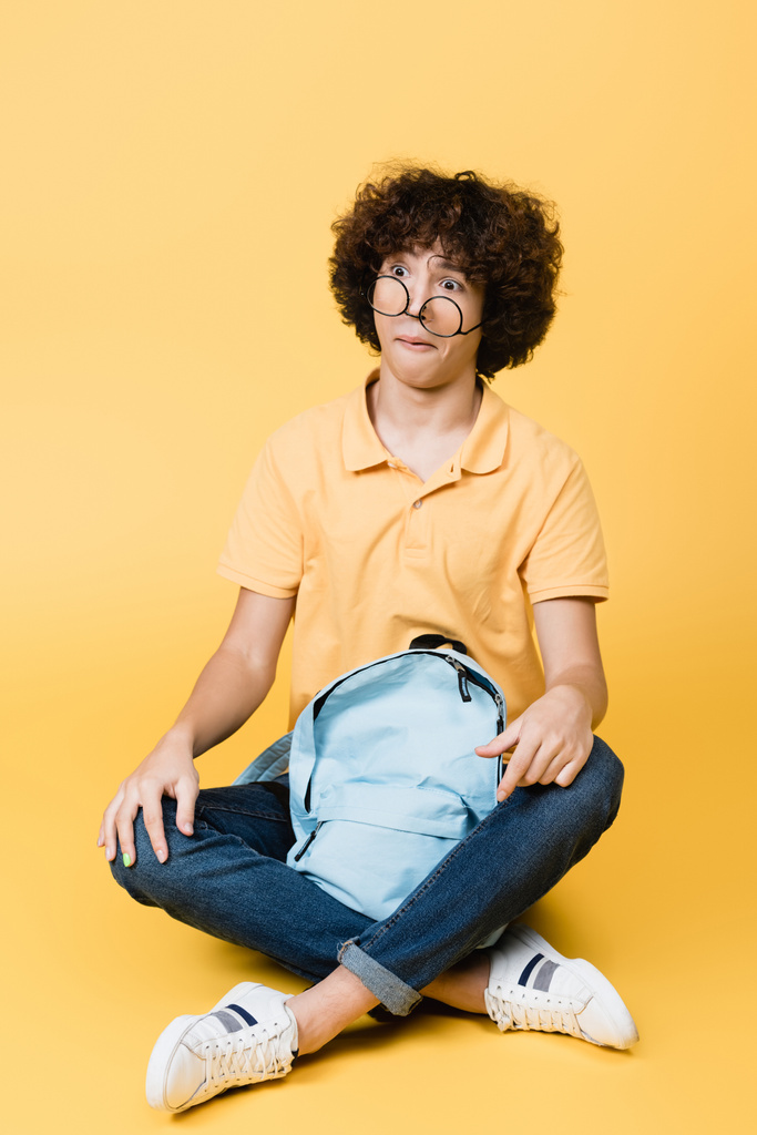 Sarı arka planda sırt çantasının yanında oturan, gözlüklü kafası karışmış kıvırcık çocuk. - Fotoğraf, Görsel