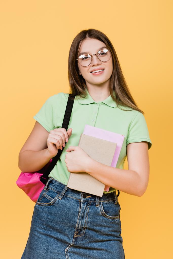 Smiling teenager in denim skirt holding notebooks and backpack isolated on yellow - Photo, Image
