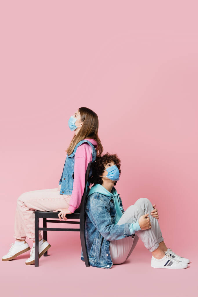 Εφηβικό αγόρι με ιατρική μάσκα κάθεται δίπλα σε φίλο στην καρέκλα σε ροζ φόντο  - Φωτογραφία, εικόνα