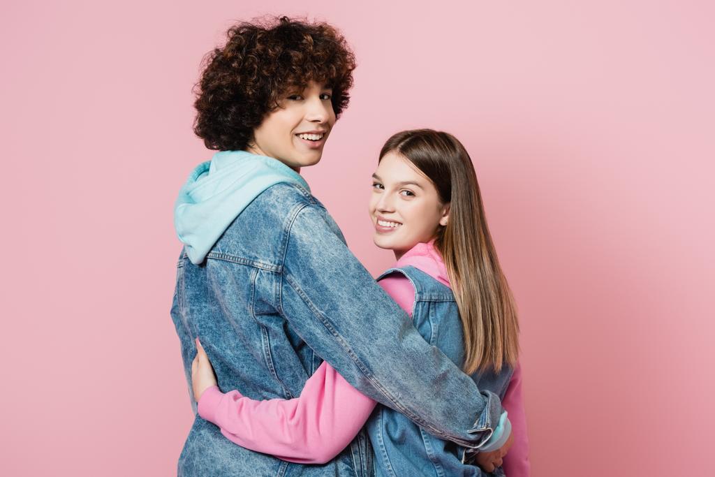 Vista posteriore degli adolescenti che sorridono alla fotocamera mentre si abbracciano isolati sul rosa  - Foto, immagini