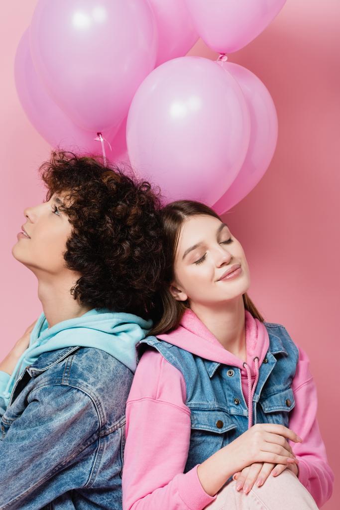 Sonriente adolescente con los ojos cerrados sentado cerca de novio y globos sobre fondo rosa  - Foto, Imagen