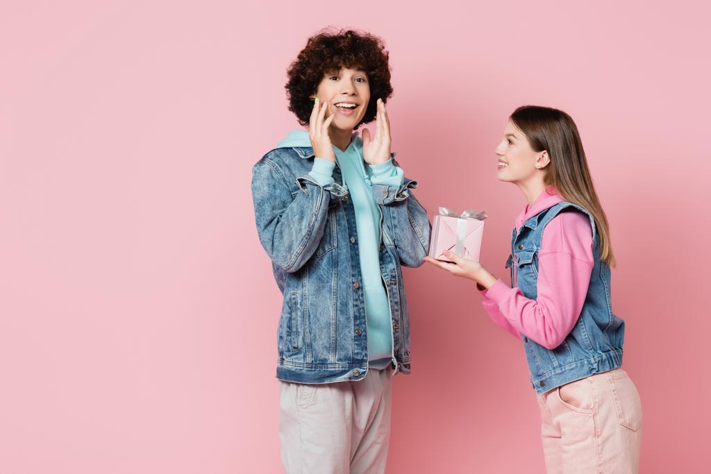 gelukkig tiener op zoek naar camera in de buurt vriendin met gift geïsoleerd op roze  - Foto, afbeelding
