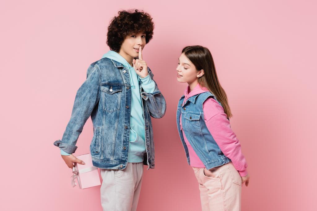 Teenager zeigt shh-Geste, während er Gegenwart in der Nähe Freundin auf rosa Hintergrund versteckt - Foto, Bild