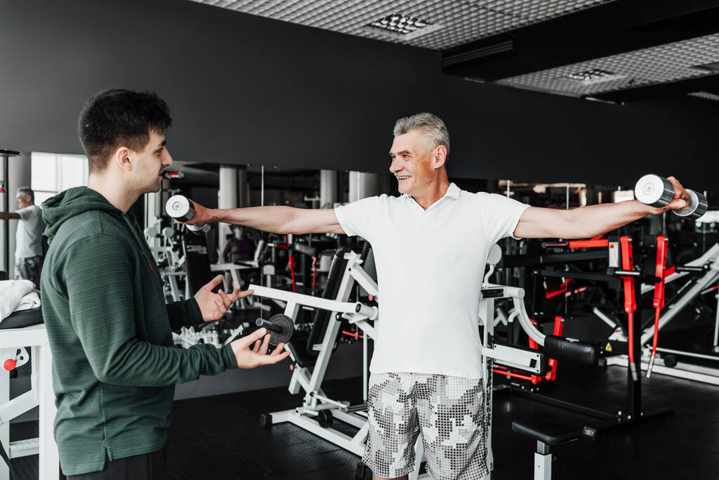 Mitten in der Turnhalle macht ein Rentner eine Übung mit Hanteln, sein Trainer beobachtet ihn und unterstützt ihn. Fitness, Sport, Gesundheit. - Foto, Bild
