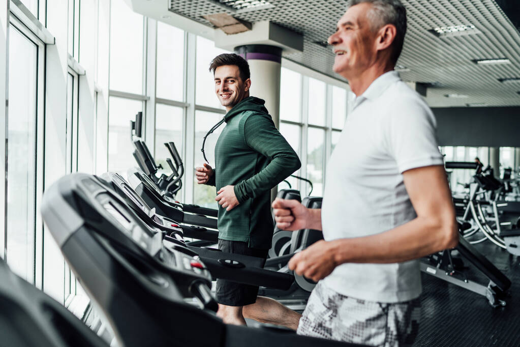 Two people on the treadmill, in the focus of a handsome young coach, he is smiling at the camera. Sports, gym, healthy lifestyle. - Photo, Image