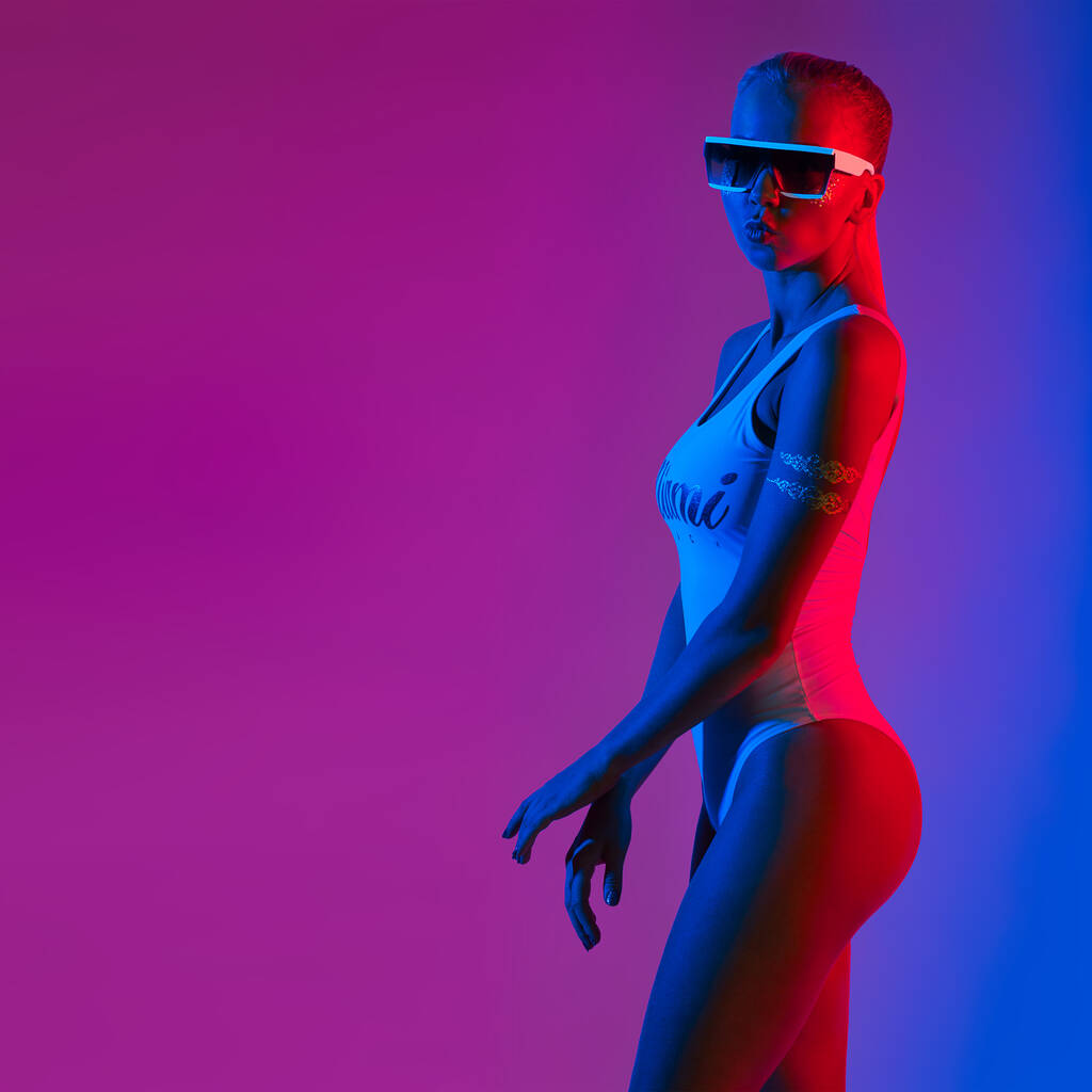 Retrato de chica joven de moda positiva en un traje de baño blanco y gafas de sol con estilo en luz de neón roja y azul y púrpura. Captura de estudio. - Foto, imagen