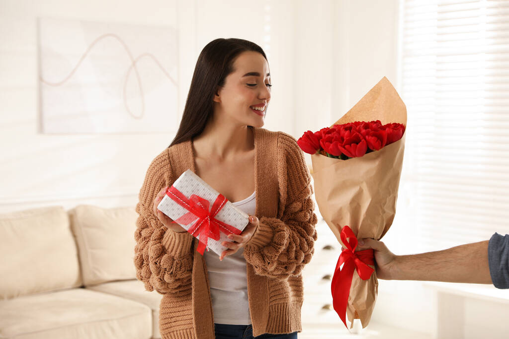 Mujer feliz recibiendo ramo de tulipán rojo y caja de regalo del hombre en casa. Celebración 8 de marzo - Foto, imagen