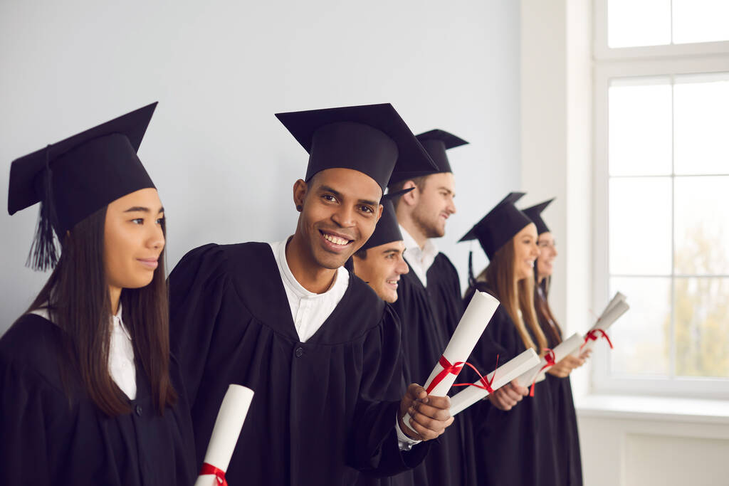 Glücklicher Absolvent mit Diplom und Blick in die Kamera, der mit anderen Studenten in einer Reihe steht - Foto, Bild