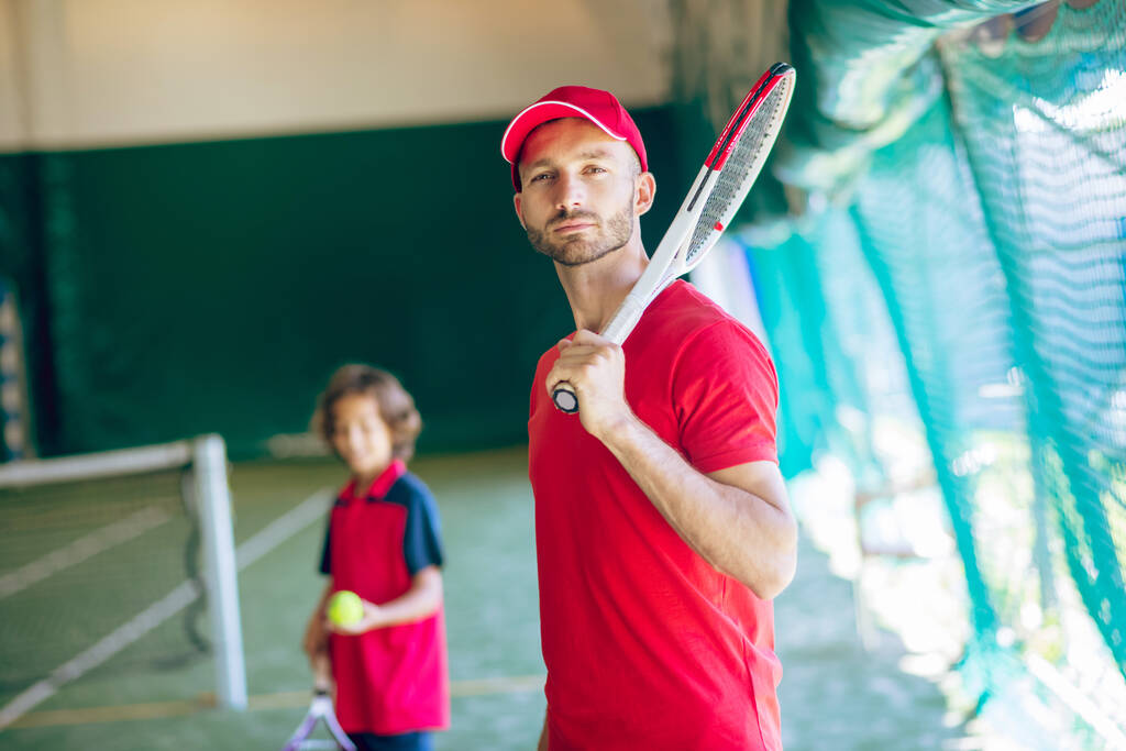 Joven barbudo con gorra roja de pie con raqueta de tenis - Foto, Imagen