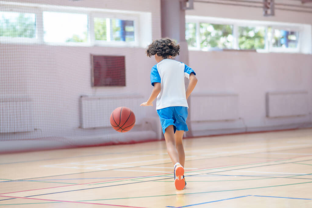Μελαχρινό αγόρι με αθλητικά να παίζει μπάσκετ σε γυμναστήριο. - Φωτογραφία, εικόνα