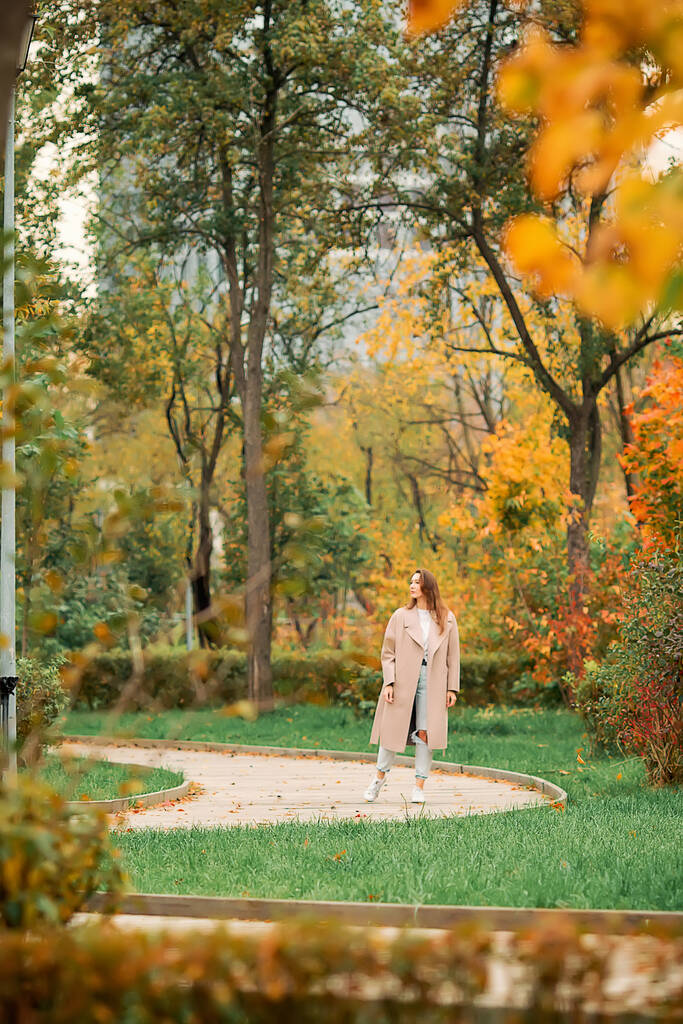 Egy fehér lány rózsaszín kabátban és kék farmerban, lyukas tánccal egy őszi parkban. - Fotó, kép