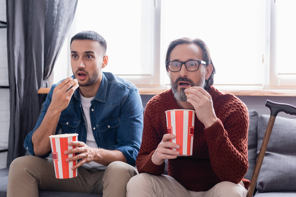 zdumiony międzyrasowy ojciec i syn jedząc popcorn podczas oglądania telewizji w domu - Zdjęcie, obraz