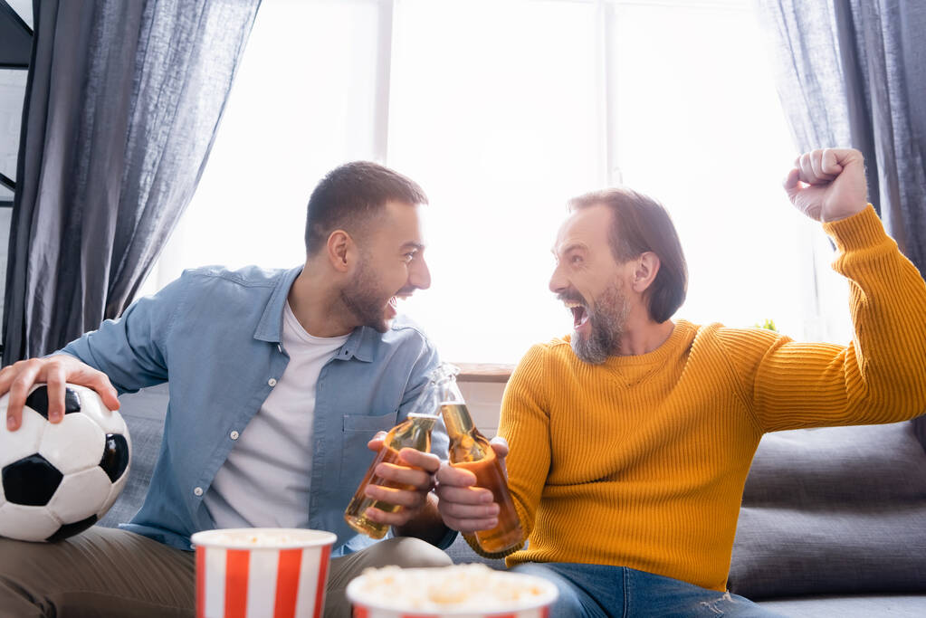 aufgeregter interrassischer Vater und Sohn klappern Bierflaschen, während sie zu Hause die Fußballmeisterschaft verfolgen - Foto, Bild