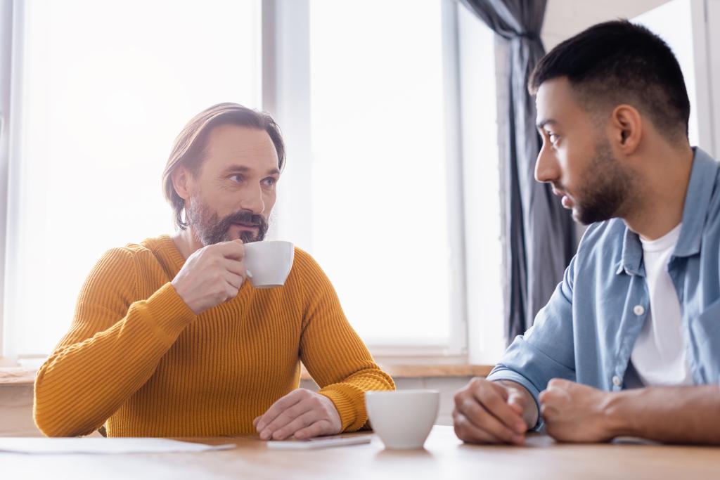 brodaty mężczyzna trzyma filiżankę kawy podczas rozmowy z hiszpańskim synem w kuchni - Zdjęcie, obraz