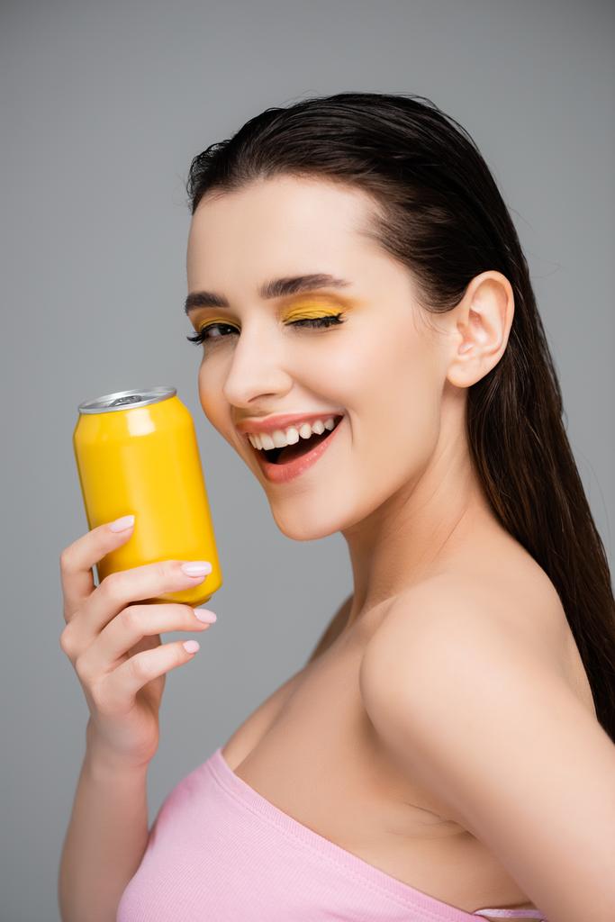 возбужденная молодая женщина держит желтую банку с безалкогольным напитком и подмигивая глаза изолированы на серый  - Фото, изображение