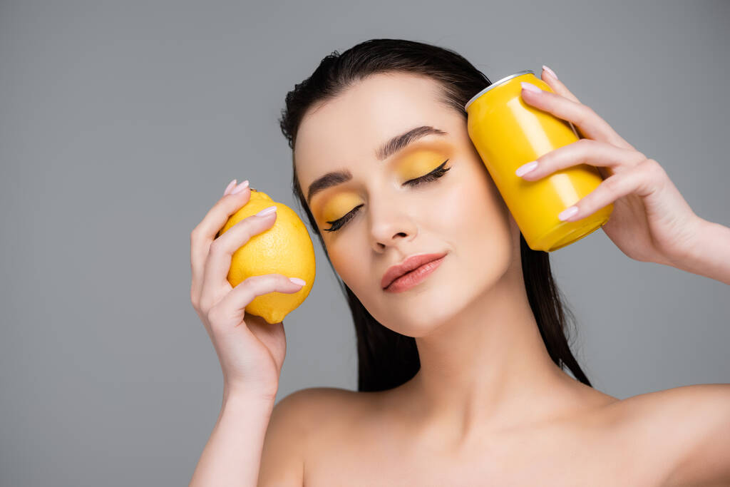 брюнетка молода жінка з закритими очима тримає жовті банки з безалкогольним напоєм і лимоном ізольовані на сірому
  - Фото, зображення
